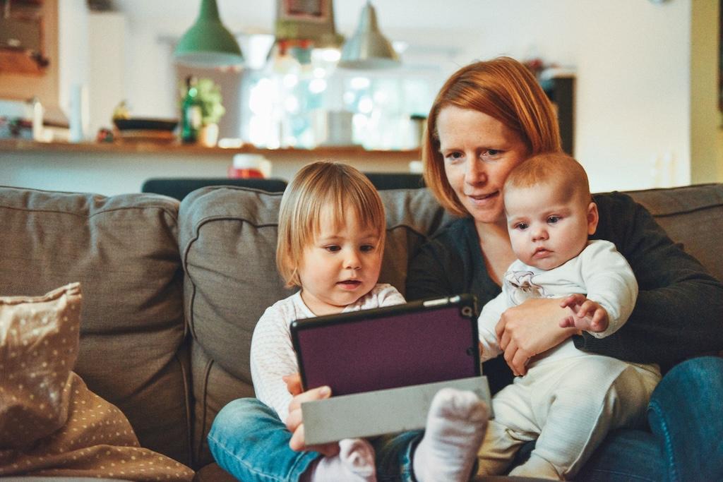 Eine Frau und zwei Kinder sitzen auf einer Couch und schauen auf ein Tablet.